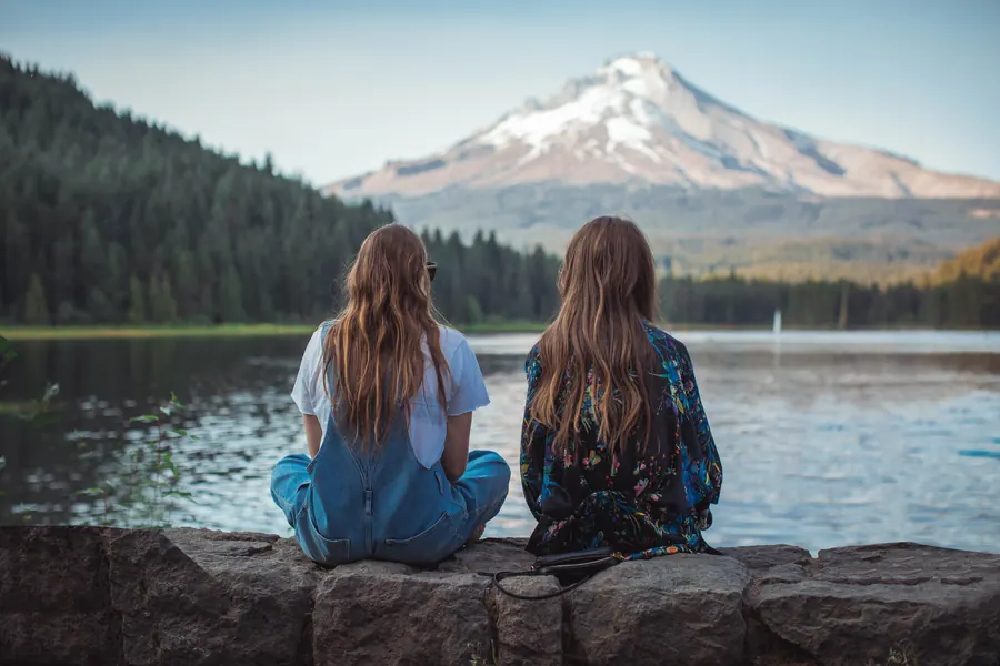 Two young women sitting by a lake, looking out at a snow-capped mountain